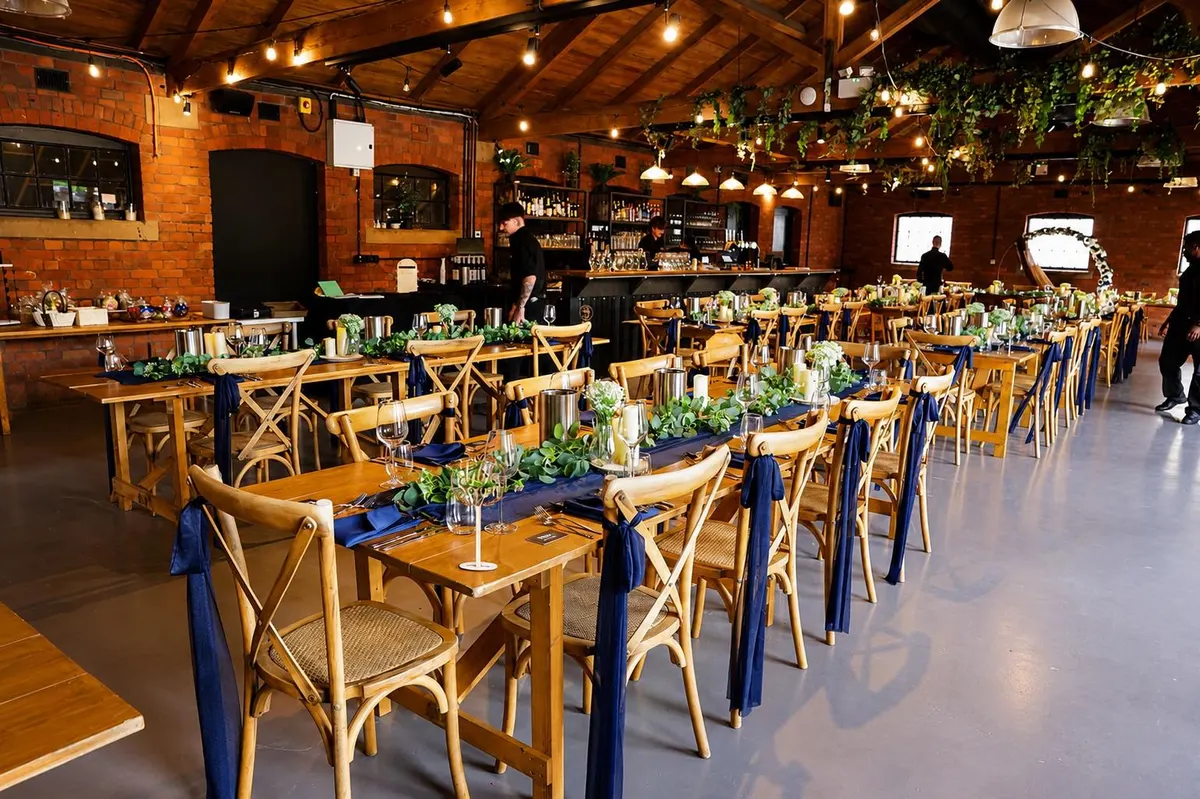 A brick-and-beam Manchester venue laid for a wedding breakfast — long trestle tables, navy runners and chair sashes, eucalyptus garlands, candles and festoon lighting.