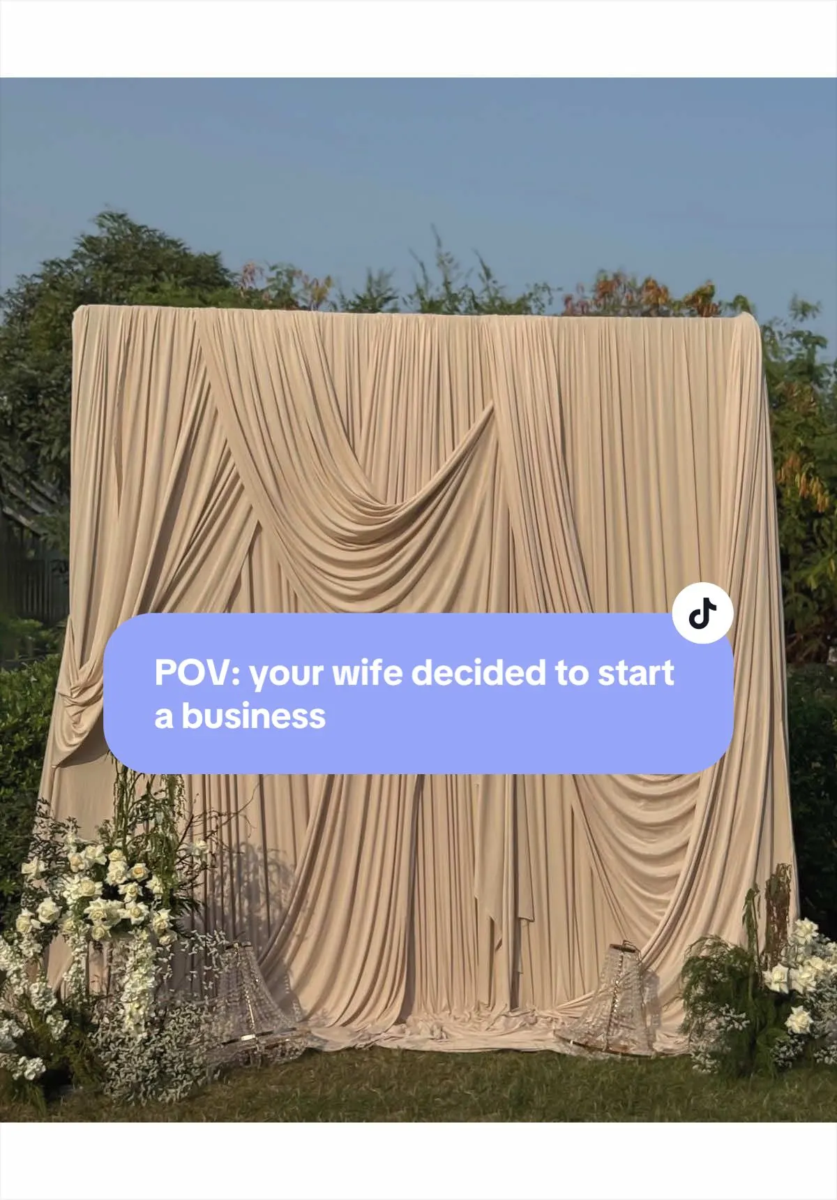 A tall champagne-coloured draped fabric backdrop set on grass under open sky, flanked by white floral and gypsophila arrangements.
