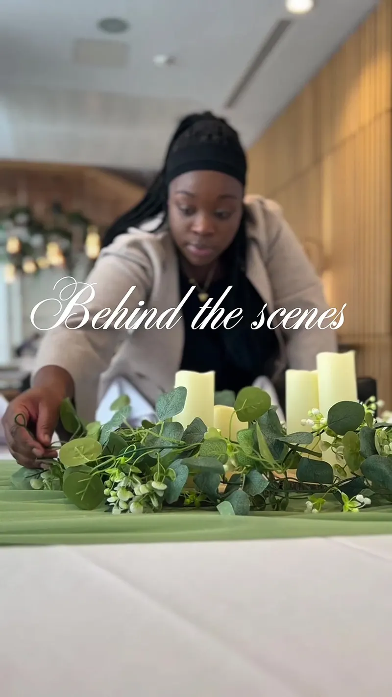 A sage green table runner dressed with eucalyptus, white blossom and ivory pillar candles being arranged by hand.