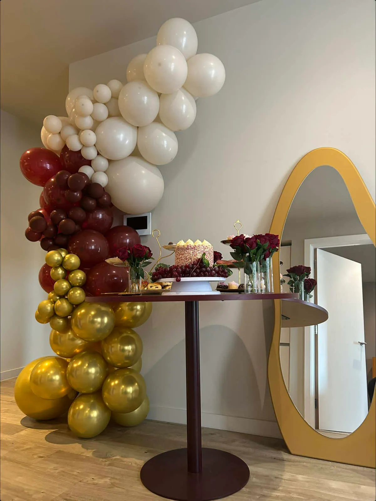 A burgundy, cream and gold balloon garland arching over a cake table with red roses, a gold-framed arch mirror and a decorated birthday cake.
