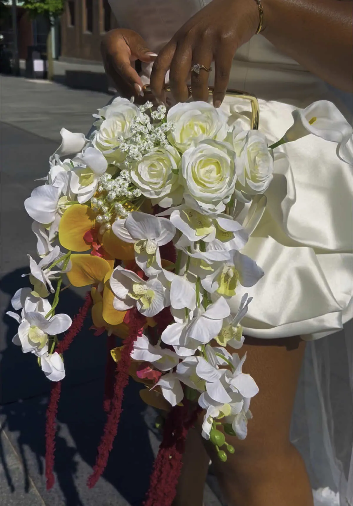 Close view of a bag bouquet of white roses, phalaenopsis orchids, amber orchids and trailing crimson amaranthus held in the sunlight.