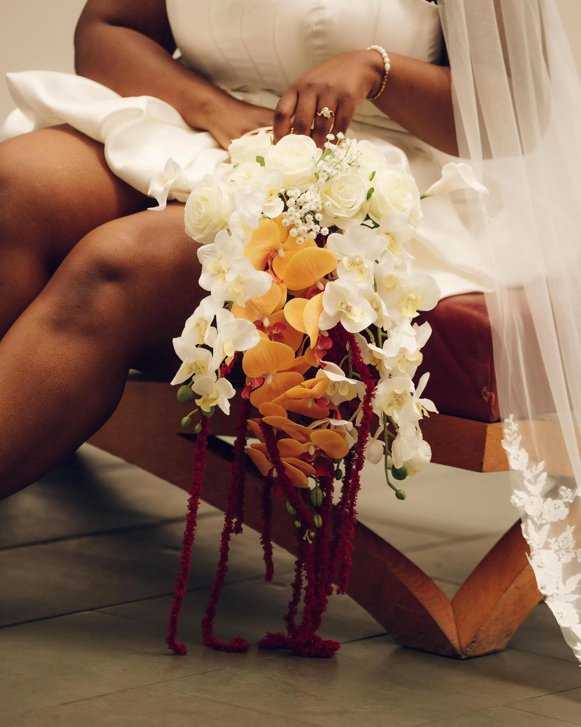 A bride seated in ivory satin holding a cascading bouquet of white roses and orchids with amber orchids and trailing crimson amaranthus, built into a white Cult Gaia bag.