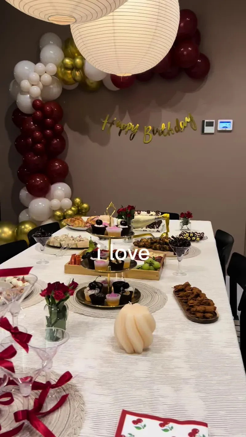 A birthday dinner table in white linen with tiered dessert stands, red roses, a sculptural candle and a burgundy, gold and white balloon garland behind.